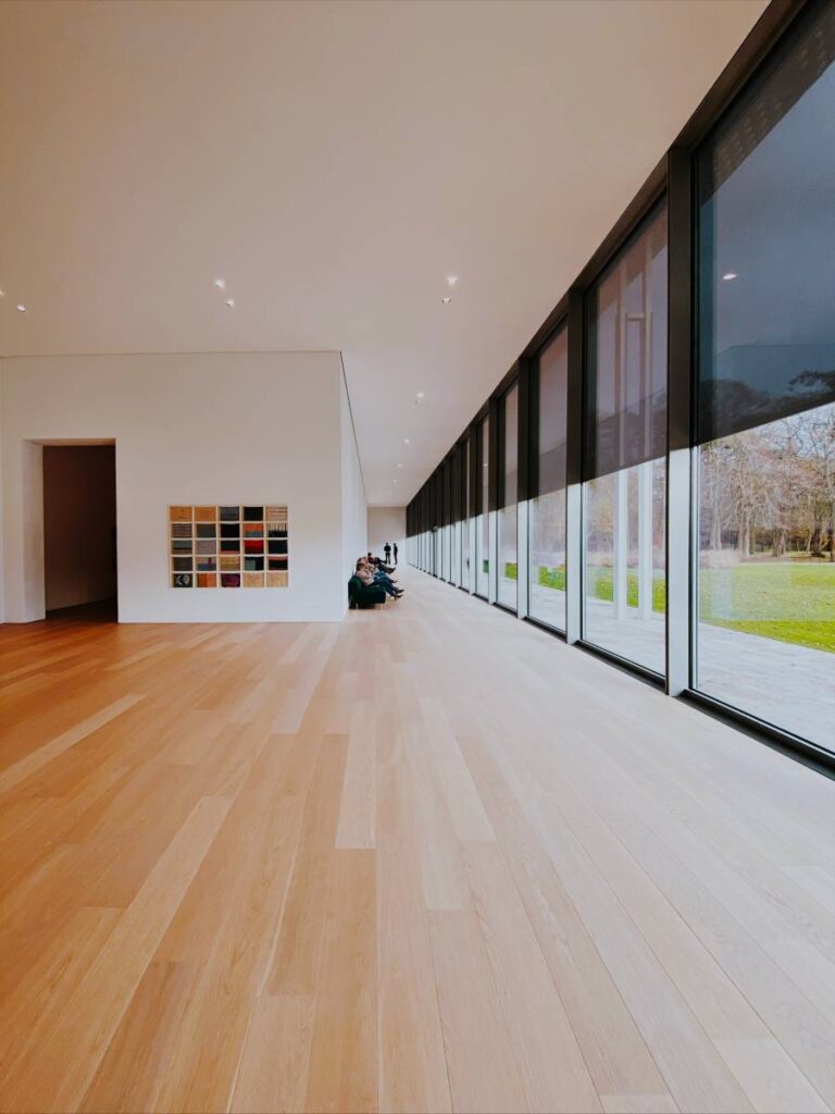 Long couloir moderne avec plancher en bois clair, grandes fenêtres sur la nature, et des personnes assises. Œuvre d'art colorée sur mur blanc.