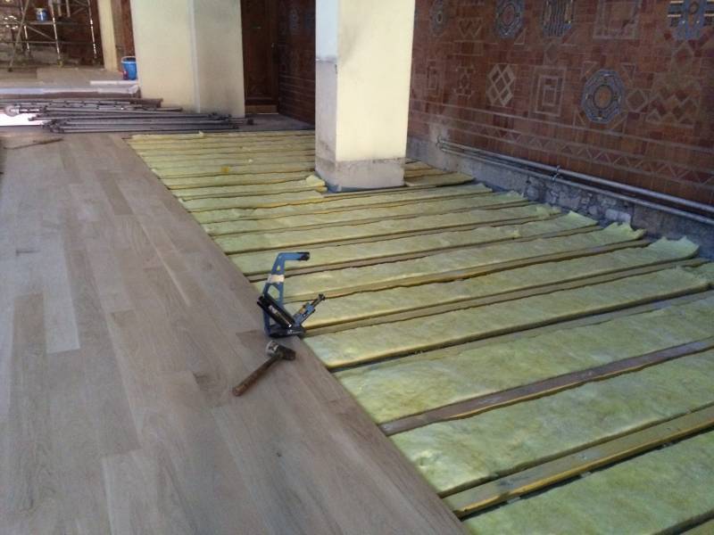 Installation de parquet clair sur un sol avec isolation jaune visible entre les lattes de bois. Un pistolet à clous et un marteau sont posés.