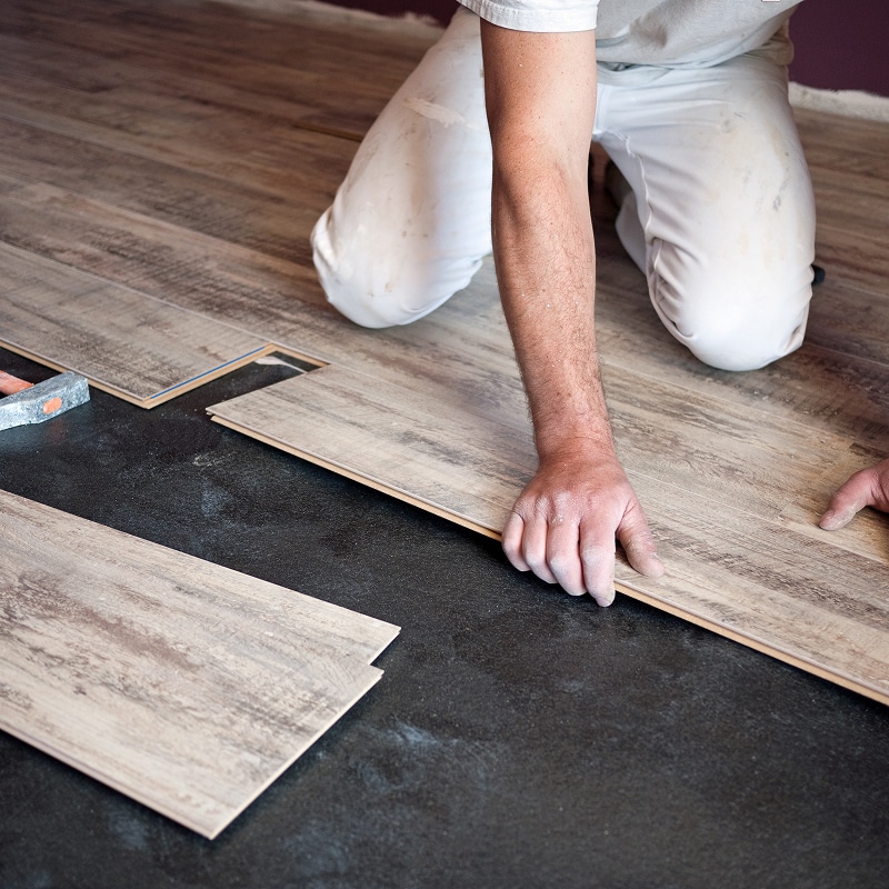 Personne à genoux installant des lames de parquet stratifié sur un sol sombre. Pose manuelle d'un nouveau revêtement de sol flottant.