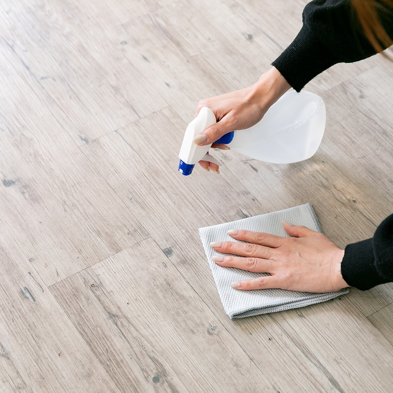 Une personne nettoie un plancher en bois clair avec un vaporisateur et un chiffon gris pour enlever la saleté.