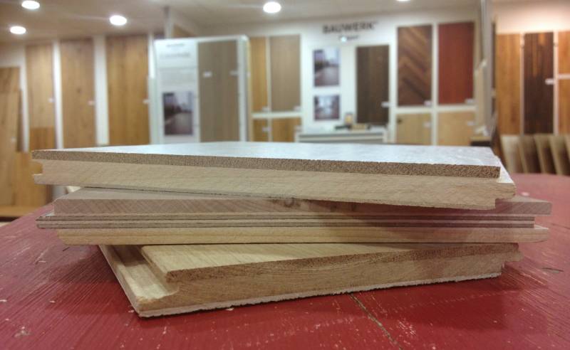 Échantillons de parquet empilés sur table rouge, bois massif et contrecollé rainuré. Showroom 'BAUWERK' en arrière-plan avec panneaux de sol.