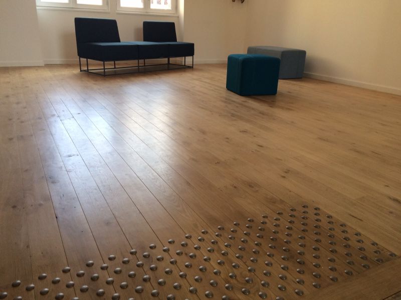 Salle lumineuse avec plancher en bois clair, deux fauteuils modulaires bleus et deux ottomans. Deux bandes podotactiles au sol.
