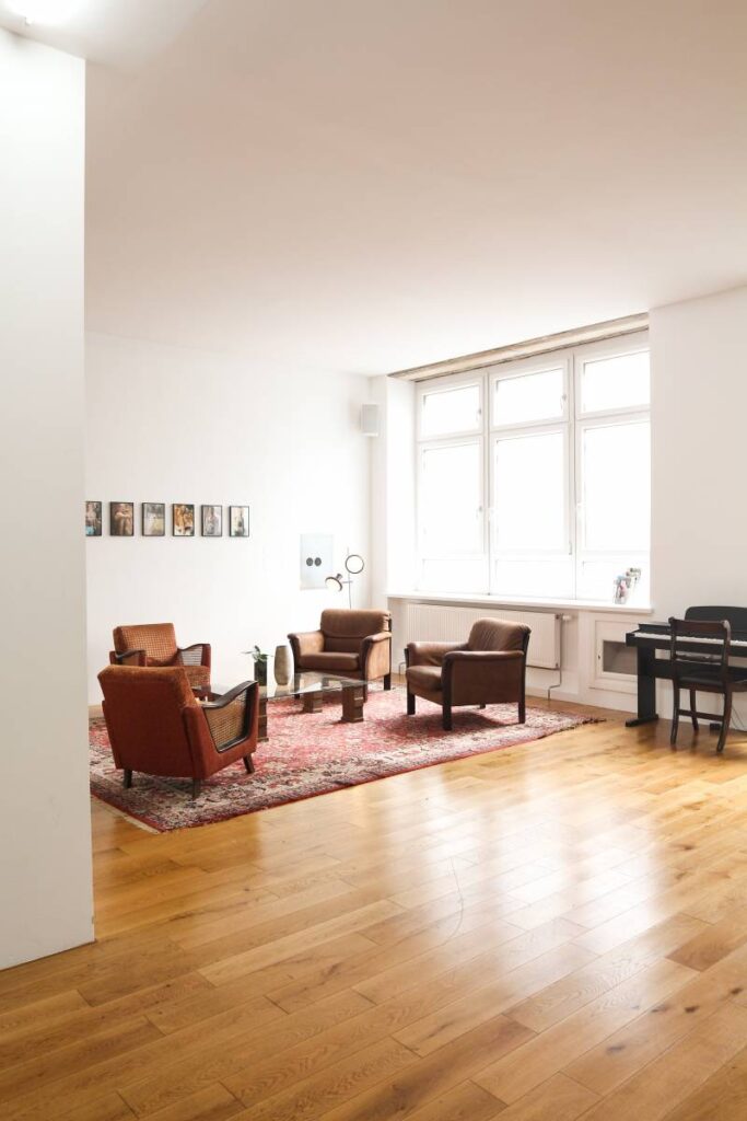 Grand salon lumineux avec parquet clair. Fauteuils vintage marron/orange sur tapis rouge, table basse, grande fenêtre et piano.
