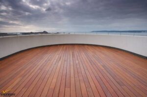 Grande terrasse en bois courbée offrant une vue panoramique sur la mer et le ciel nuageux, avec un parapet blanc moderne.