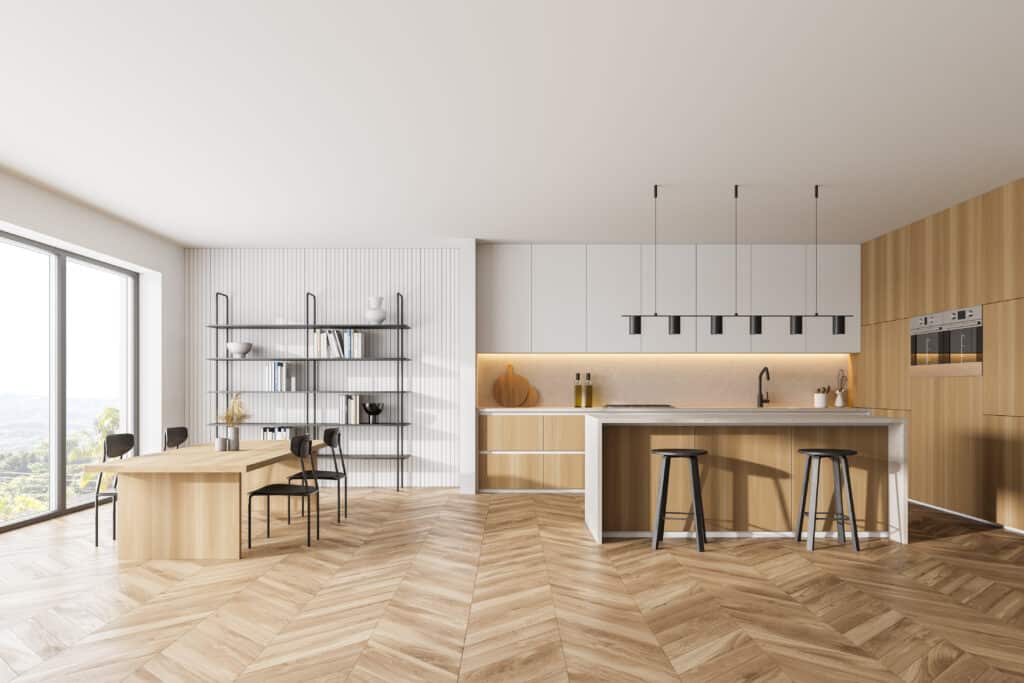 Cuisine moderne ouverte bois clair et blanc, îlot central, salle à manger avec table en bois et chaises noires. Parquet chevrons, vue nature.