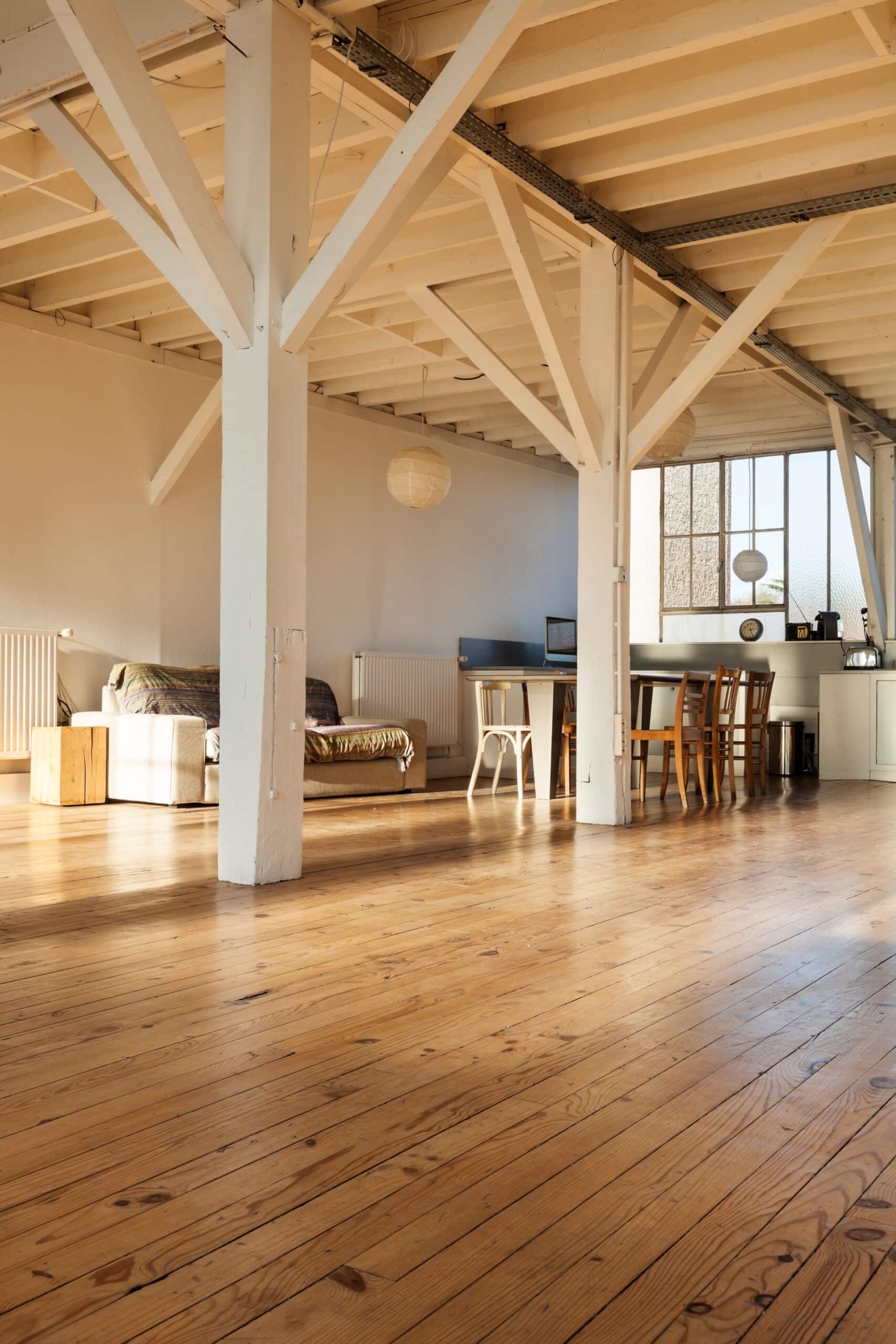 Grand loft lumineux avec poutres et colonnes en bois blanc, plancher en bois clair, salon, salle à manger et grandes fenêtres.