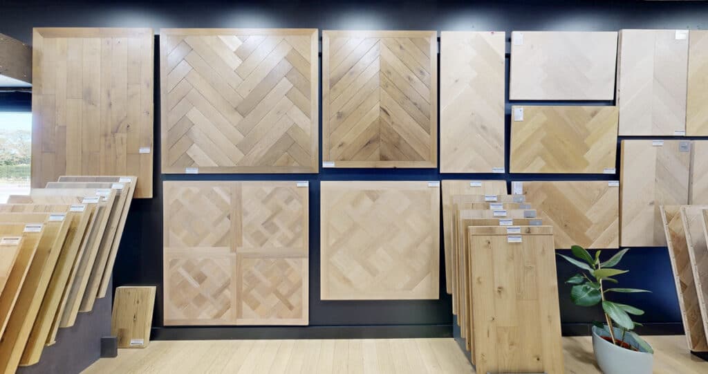 Showroom de parquets en bois clair: grands échantillons muraux (chevrons, lames droites, motifs variés) et dalles sur présentoirs. Plante verte.