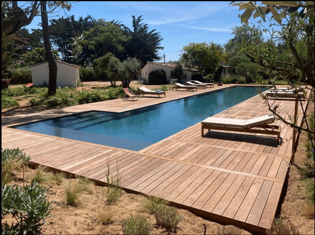 Grande piscine rectangulaire entourée d'une vaste terrasse en bois clair avec chaises longues, sous un ciel bleu estival, dans un jardin.