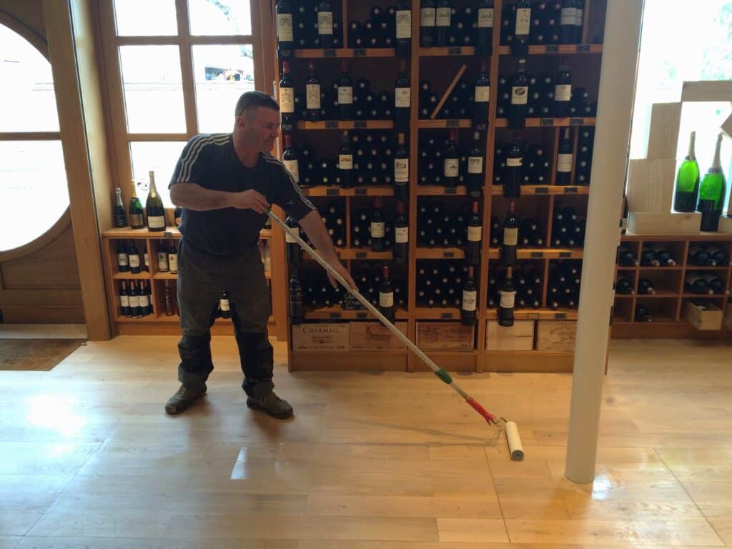 Homme appliquant du vernis sur un parquet en bois clair avec un rouleau. Au fond, un grand casier à vin rempli de bouteilles, dont des boîtes "CHARMAIL".