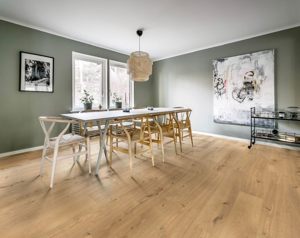 Salle à manger moderne aux murs verts, table blanche, chaises en bois clair, parquet clair. Suspension tressée, tableaux muraux.